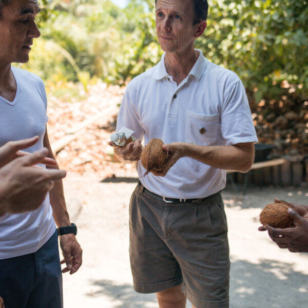 A group of people stands outdoors, engaging in conversation. One person in light shorts and a white polo shirt holds two coconuts, embodying the Maldives exclusive experience. Another person, slightly in frame, points towards the coconuts. The background is filled with greenery and a sunny atmosphere.