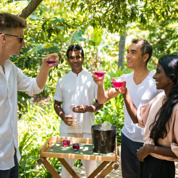 Four people stand outdoors in a lush garden, smiling and clinking glasses filled with a pink drink. Three are near a wooden table with an ice bucket, while the fourth stands behind them, holding a glass and smiling—an exclusive experience reminiscent of the serene beauty found at Soneva in the Maldives.