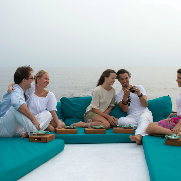 Five people sit on teal cushions on a boat, smiling and laughing. They are casually dressed, each with a tray of food. One person is showing photos on a camera. The background features the calm ocean under an overcast sky, capturing their Maldives Exclusive Experience with Soneva.