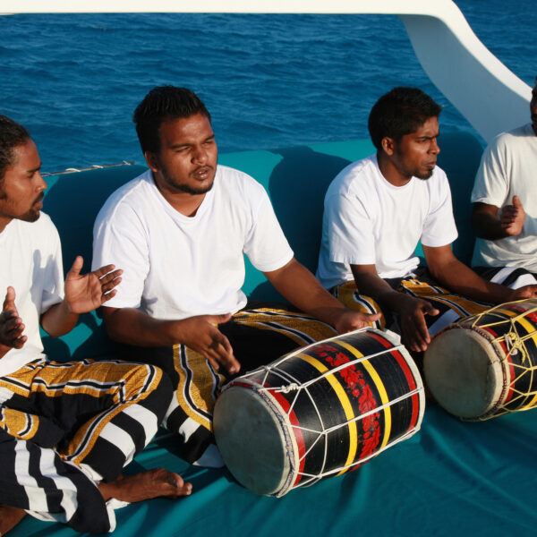 Four men are sitting on a boat in front of the blue ocean, playing traditional drums and clapping. They are all wearing white shirts and colorful, patterned pants. Experience this Maldives Exclusive at Soneva under a clear, bright sky.