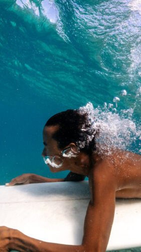 A surfer with short hair and wearing striped shorts is underwater, holding onto his surfboard as a wave passes over him. The turquoise water, part of the Maldives exclusive experience at Soneva, is clear, showing bubbles around the surfer's face and body.