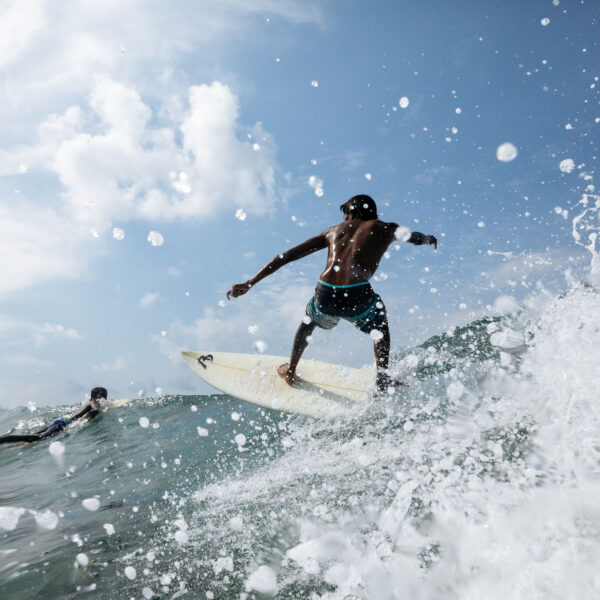 A surfer balances on a wave under a clear blue sky, surrounded by splashing water in this Maldives exclusive experience. The surfer wears a black cap, dark shorts, and a white watch. Another person is seen paddling in the background on a surfboard, embodying the serene adventure offered by Soneva resorts.