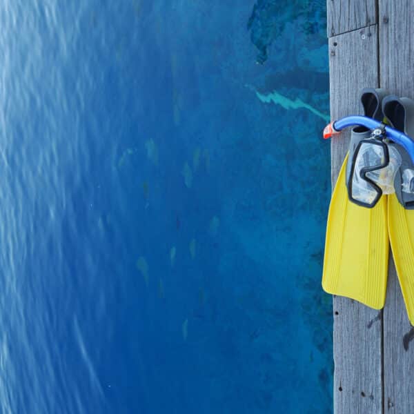 A pair of yellow flippers and a blue snorkel with a mask rest on a wooden pier above clear, blue water. The calm water shows some hints of underwater marine life near the surface, promising an exclusive experience reminiscent of Soneva resorts in the Maldives.