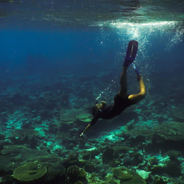 A snorkeler swims underwater in the clear blue ocean of the Maldives, surrounded by coral reefs and marine life. The sunlight filters through the water, illuminating the scene. The snorkeler extends one arm and kicks with fins, exploring this exclusive Soneva experience in a vibrant underwater landscape.