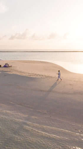 A lone figure walks on a narrow sandy beach that stretches into the tranquil sea under a clear sky. In the background, a white umbrella set up with a few people sitting beneath it provides shade from the bright sun. The vast expanse of water glistens in the sunlight, reflecting the essence of a Maldives-exclusive experience at Soneva.