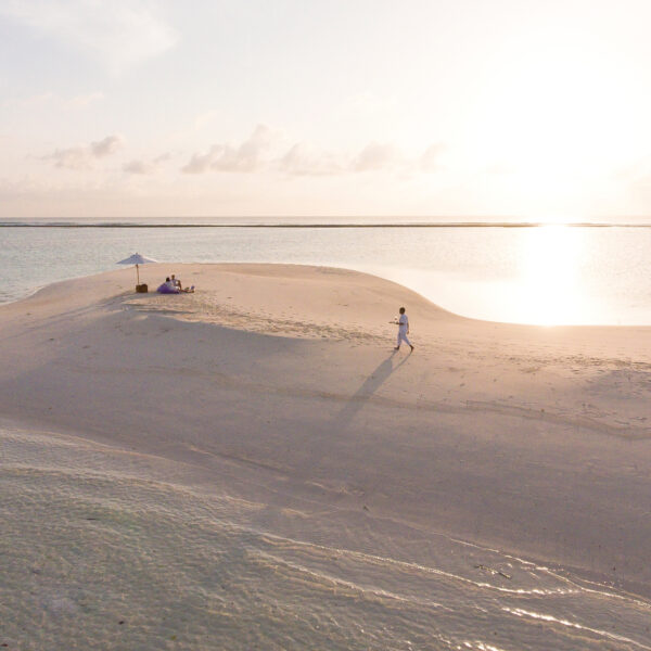 A lone figure walks on a narrow sandy beach that stretches into the tranquil sea under a clear sky. In the background, a white umbrella set up with a few people sitting beneath it provides shade from the bright sun. The vast expanse of water glistens in the sunlight, reflecting the essence of a Maldives-exclusive experience at Soneva.