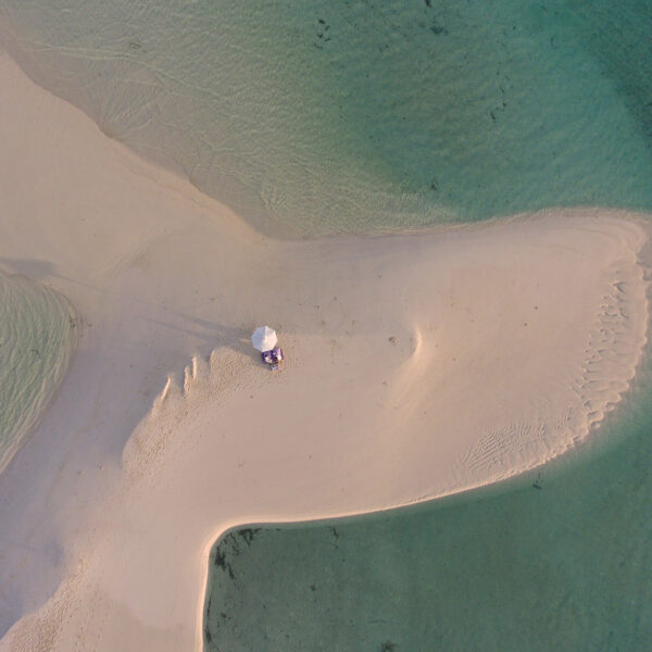 Aerial view of a person with an umbrella standing on a small, sandy peninsula in the Maldives, jutting into clear, turquoise water. The sand forms a unique shape resembling a fish tail, surrounded by the calm sea. This exclusive experience at Soneva reveals visible shoreline and sand textures.