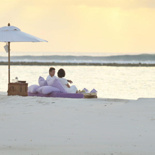 A couple sits on a cozy setup with purple cushions and a small table under a white umbrella on a serene beach at sunset. The calm ocean stretches out to the horizon. This Maldives exclusive experience, offered by Soneva, is both peaceful and romantic.