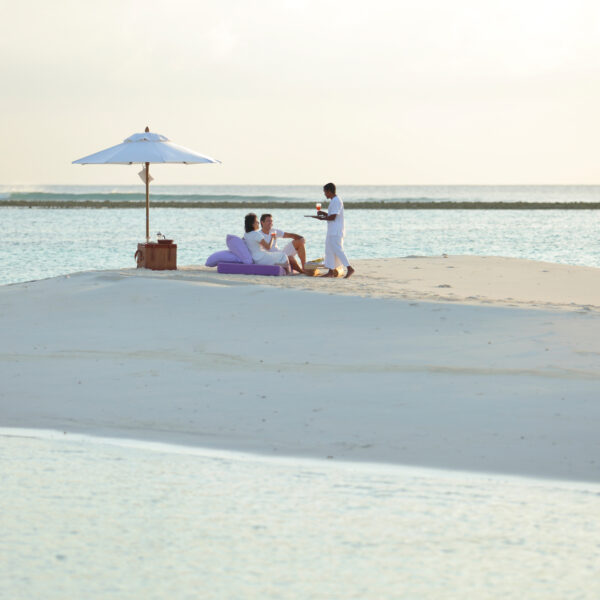 Two people relax under a white umbrella on a small sandy island surrounded by clear blue water in the Maldives. They sit on purple cushions, enjoying refreshments served by a person in white. The scene is serene, with the horizon and gentle waves in the background—a true Soneva exclusive experience.