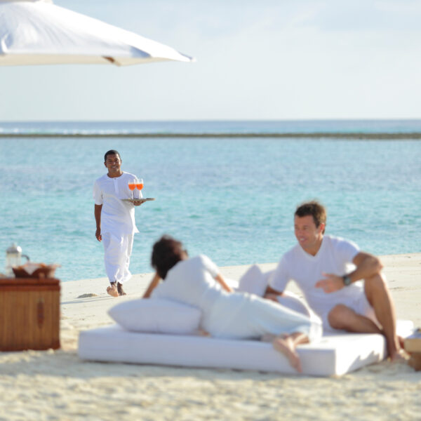 A couple relaxes on a beach, lying on white cushions under a large white umbrella. A server dressed in white approaches them with a tray holding two drinks. The ocean and a clear sky are visible in the background, creating a serene and luxurious atmosphere as part of the Maldives Exclusive Experience at Soneva.