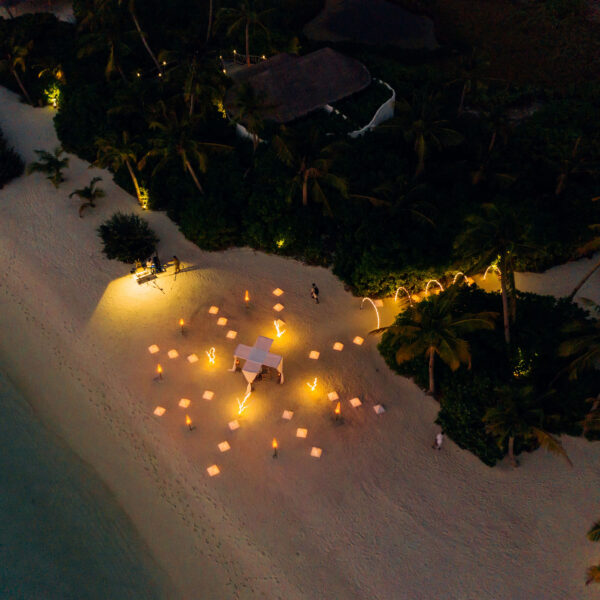 Aerial view of a nighttime beach scene with glowing lanterns arranged in clusters on the sand. Palm trees and dense greenery border the beach, and a small structure with a roof is nestled among the trees. Soft light illuminates parts of the trees and sandy shore, creating a Maldives exclusive experience.