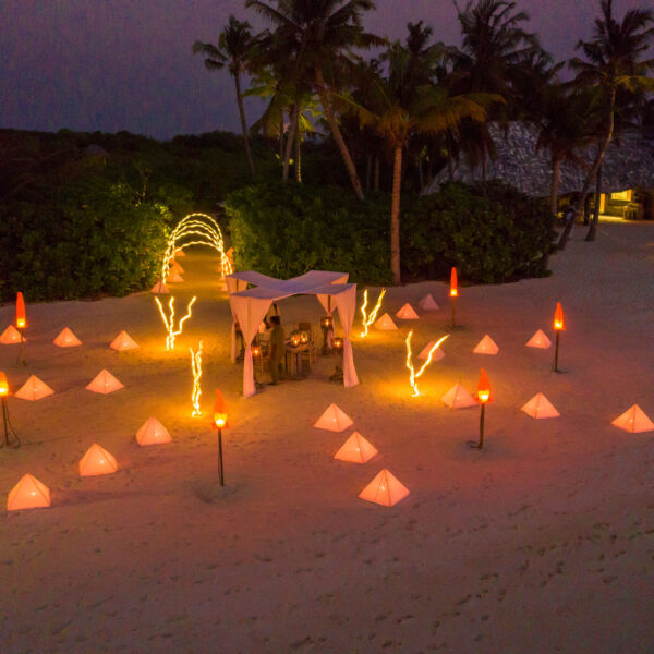 An aerial view of an illuminated beach setup at dusk reveals a Maldives exclusive experience. A central gazebo is surrounded by glowing pyramid-shaped lights arranged in a circle. Fairy lights and torches add to the ambiance, with palm trees and buildings in the background.