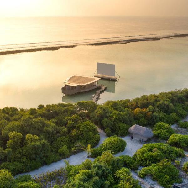 A tranquil aerial view shows a rustic, open-air cinema on stilts over the clear waters of the Maldives. Accessible by a pier and surrounded by lush greenery, this Soneva exclusive experience reveals the sea stretching into the horizon under a warm, setting sun.