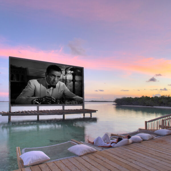 Outdoor movie screening on a floating platform in a serene, tropical setting. The black-and-white film is displayed over calm water, surrounded by a wooden dock with cushions for seating. As the sun sets, painting the sky in soft pinks and purples, indulge in this Maldives exclusive experience at Soneva.