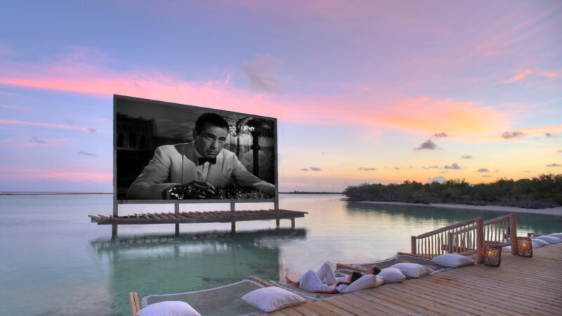 Outdoor movie screening on a floating platform in a serene, tropical setting. The black-and-white film is displayed over calm water, surrounded by a wooden dock with cushions for seating. As the sun sets, painting the sky in soft pinks and purples, indulge in this Maldives exclusive experience at Soneva.