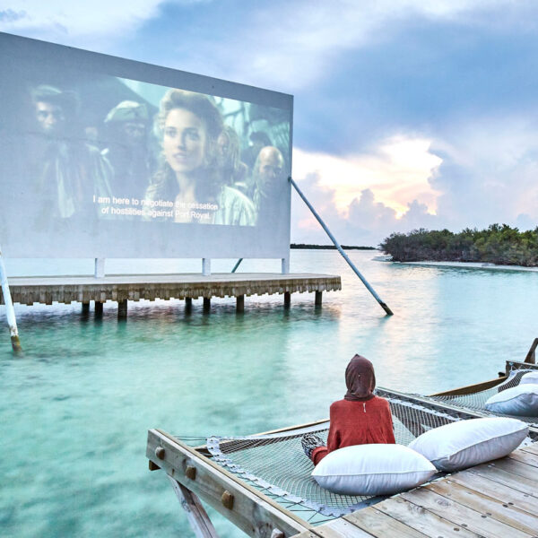 An outdoor floating cinema screen displays a movie over calm, turquoise waters in the Maldives. In the foreground, a person sits on a wooden deck on a netted hammock with white pillows, watching the screen. The sky is cloudy with hints of dawn or dusk light, capturing an exclusive experience at Soneva.