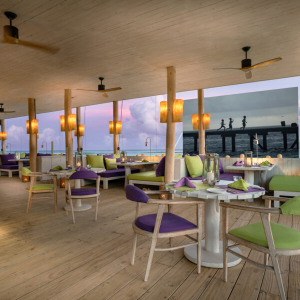 A cozy, open-air restaurant with wooden floors and a wooden ceiling. The dining area features green and purple cushioned seating, round tables, and hanging yellow lanterns. Enjoy a Maldives Exclusive Experience as the sunset view through large windows enhances the ambiance with soft, warm lighting.