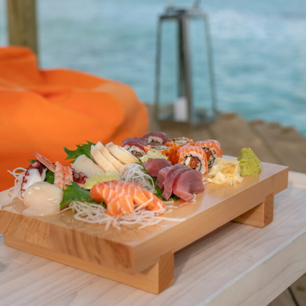 A wooden tray filled with assorted sushi, sashimi, and garnishes sits on a light wooden table. An orange bean bag and a blurred view of a body of water are visible in the background, suggesting a relaxed, outdoor dining setting that's part of the exclusive Soneva experience in the Maldives.