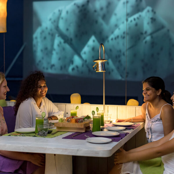 Four women are sitting around a rectangular dining table outdoors, enjoying a meal together. The table has green drinks, plates, and appetizers on it. The scene is lit by a lamp above the table, and a large screen in the background displays an image of dragon fruit – the perfect Maldives Exclusive Experience at Soneva.