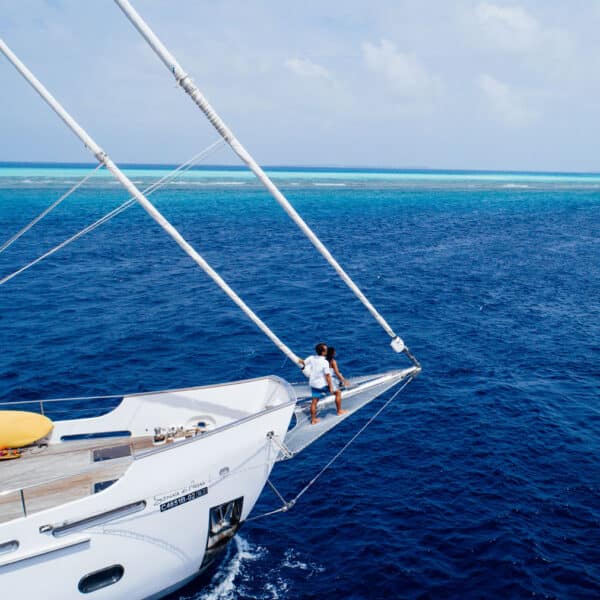 A person stands at the bow of a sailboat cruising through the deep blue waters under a clear Maldivian sky. They are holding onto the rigging. A yellow lifeboat is visible on the deck. The horizon shows a strip of turquoise water, indicating a nearby reef, embodying the essence of a Soneva exclusive experience.