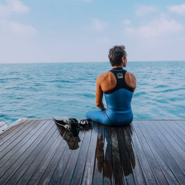 Person in a blue wetsuit sits on a wooden deck overlooking a calm, expansive ocean. A pair of flippers rests beside them, and the sky is clear with a few clouds. The person appears to be enjoying a tranquil moment as part of their Maldives exclusive experience at Soneva.