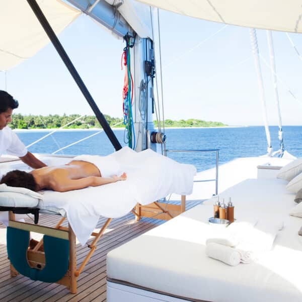 A person receives a massage on a luxury yacht's deck under a canopy during their Maldives holiday. The massage therapist, wearing white, stands beside the table. Cushioned seating, towels, and a serene ocean with distant greenery add to the tranquil ambiance. The sky is bright and clear.