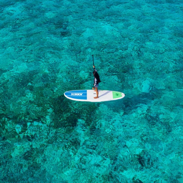 A person stands on a paddleboard over clear turquoise waters, surrounded by visible underwater rocks and coral at the Maldives, offering an exclusive Soneva experience. The individual is seen from an aerial perspective, paddling through the serene, transparent ocean.