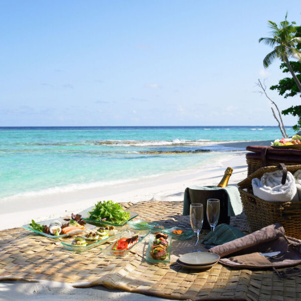 A romantic beach picnic setup with a wicker basket, champagne bottle, flutes, towels, and a variety of fresh food on a woven mat. The scene overlooks turquoise waters, white sand, and lush greenery under a clear blue sky—truly a Maldives Exclusive Experience by Soneva.