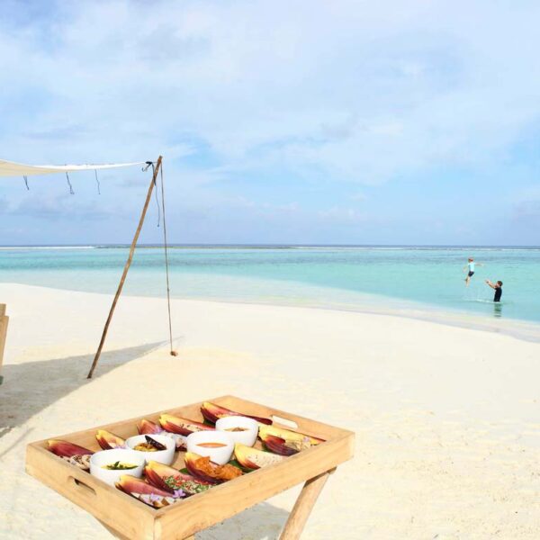 A beautiful beach scene with a wooden table and chairs set under a canopy on the white sand. A tray with assorted food is in the foreground. In the background, a person stands in the shallow turquoise water, and another person sits on the sand near the shoreline, enjoying this exclusive Maldives experience at Soneva.