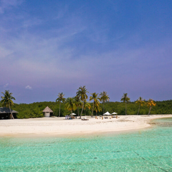 A tropical beach scene with clear turquoise waters, white sandy shore, and a backdrop of lush green vegetation and palm trees. Soneva offers an exclusive Maldives experience with several thatched-roof structures and lounge chairs with umbrellas scattered along the beach under a clear blue sky.