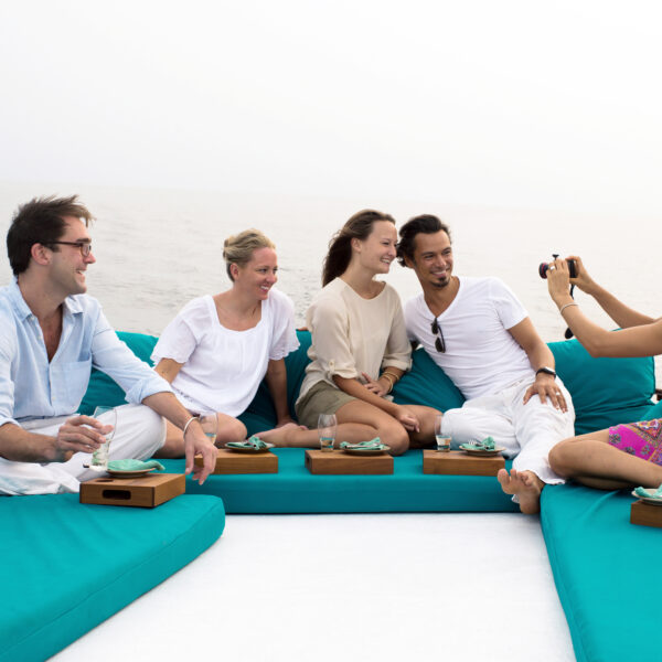 Five people enjoying a Maldives exclusive experience on the deck of a boat, sitting on turquoise cushions with the ocean backdrop. Four of them, two men and two women, smile and pose for the camera as a fifth person, a woman, captures this Soneva moment.