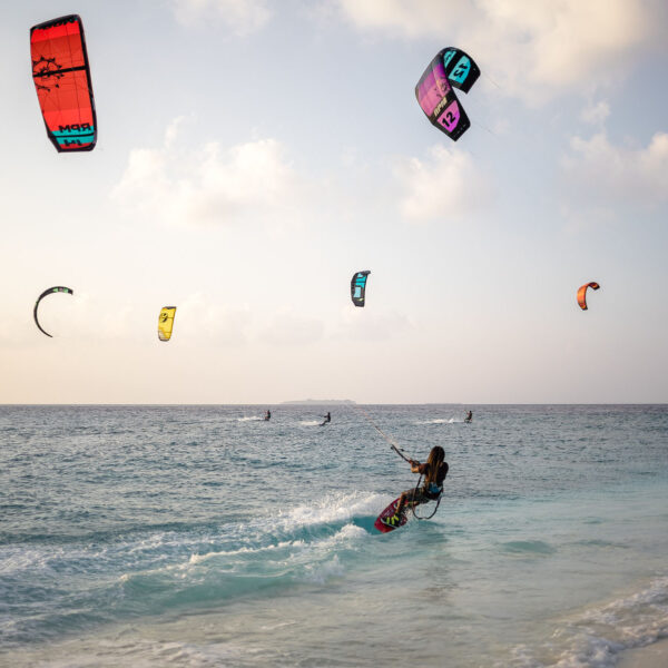 A group of kite surfers are riding the waves near a sandy shore at Soneva in the Maldives, with brightly colored kites soaring in the sky against a backdrop of blue ocean and a partly cloudy sky. The scene captures the excitement and motion of this exclusive experience.