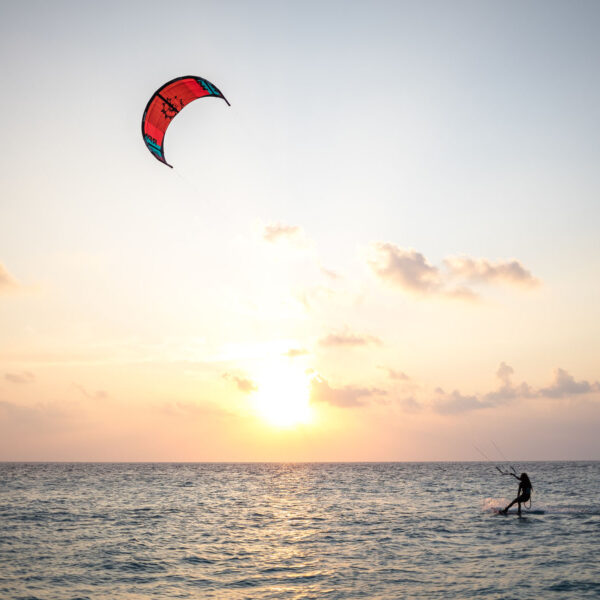 A person is kiteboarding on the ocean at sunset, enjoying an exclusive experience in the Maldives. The sky is painted with soft hues of orange and yellow, with a few scattered clouds. The red-and-black kite soars high as the rider glides smoothly across the waves near Soneva resort.