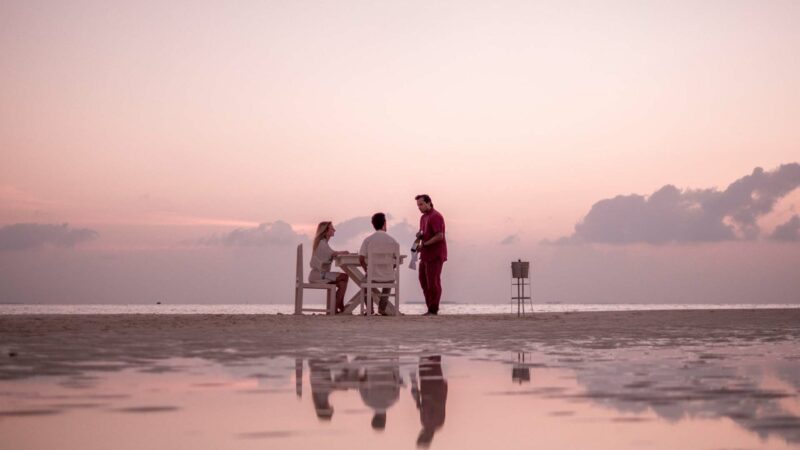 Three people relax at a beach during sunset. Two are seated at a table while another stands beside them, perhaps discussing their recent Maldives luxury yacht holiday. The sky is pinkish-purple, reflecting off the wet sand and creating a serene ambiance. Clouds are dark, contrasting with the soft colors of the horizon.