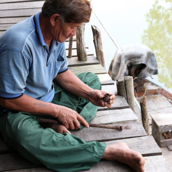 A person sits on a wooden pier by the water, handling a small object. Wearing a blue shirt and green pants, they are surrounded by various tools and objects. This scene captures an exclusive experience in Thailand at Soneva that&#039;s both serene and immersive.