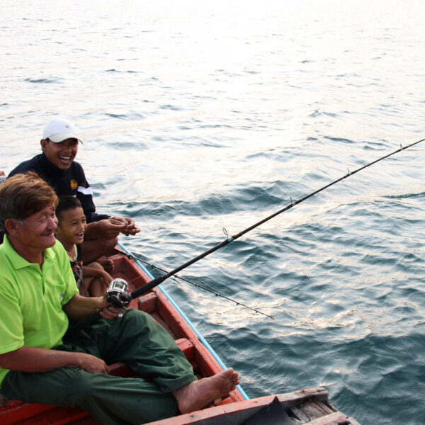 A group of four smiling people sit on the edge of a wooden boat in Thailand. One person in a green shirt is holding a fishing rod with a small fish on the line. The boat is on calm water, and they all appear to be enjoying this exclusive Soneva experience.