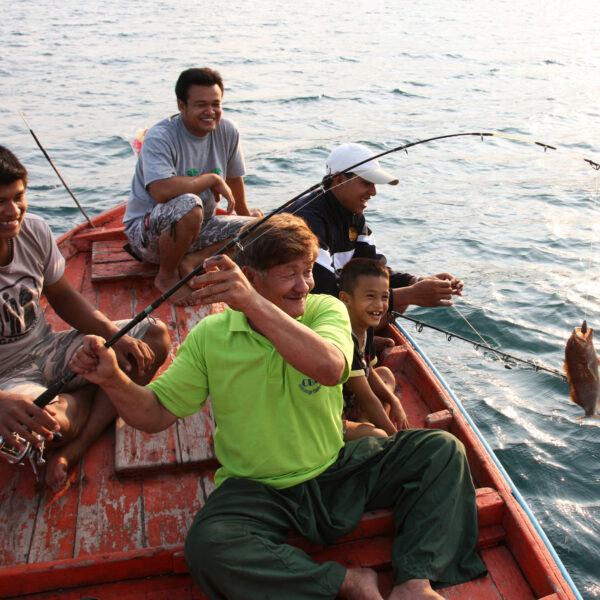 Five people are sitting on a wooden boat in the water, enjoying a fishing trip. One of them, a man wearing a green shirt, is reeling in a fish with a fishing rod. Everyone appears to be smiling and having a good time with an ocean view surrounding them—truly, it&#039;s an exclusive Soneva experience in Thailand.