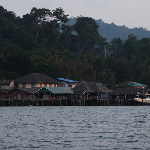 A coastal village with wooden houses by the water, surrounded by lush green trees and hills in the background. A boat is docked by the village, and the sky above is overcast—a perfect Thailand exclusive experience that promises tranquility and natural beauty.