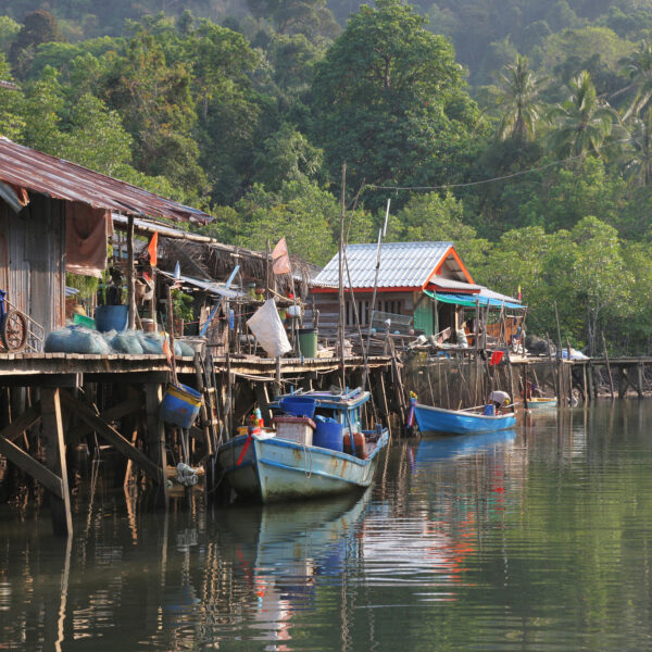 Discover a serene fishing village in Thailand, where wooden houses on stilts stand over calm waters. Colorful boats are moored along the shore, and lush green trees provide a natural backdrop. A wooden walkway connects the homes, while fishing nets and equipment are visible around this exclusive experience.