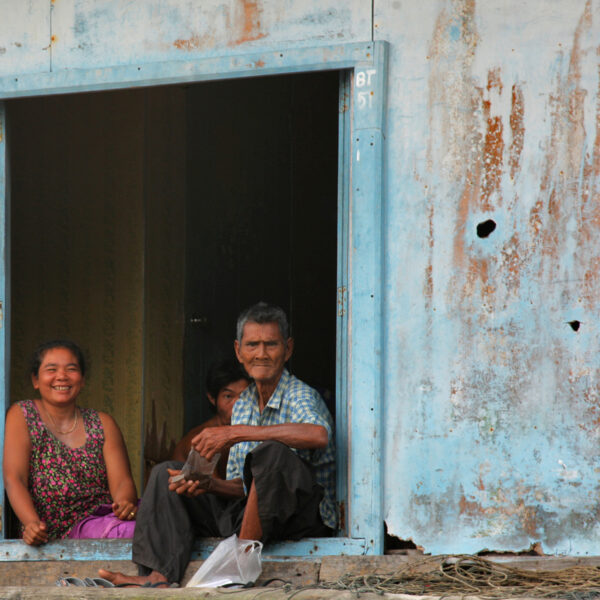 A smiling woman, a man, and a child sit in the doorway of a weathered blue building. The exterior appears rustic with peeled paint and patches of rust, reminiscent of an exclusive experience at Soneva in Thailand. The woman wears a colorful dress, and the man holds something in his hands.