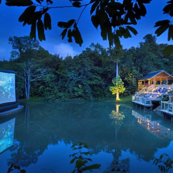 Outdoor movie theater at twilight with a screen set up on a platform over a serene pond, surrounded by lush trees. On the right, tiered seating with wooden structures and purple cushions offers a clear view of the screen. This Thailand exclusive experience at Soneva is tranquil and picturesque.