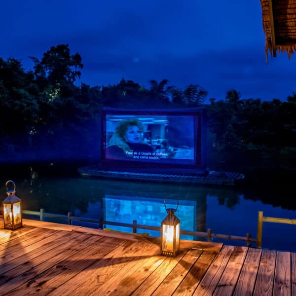 An outdoor movie screen is set up over a calm body of water at dusk, surrounded by trees. Two lit lanterns rest on a wooden deck in the foreground, casting a warm glow. This Thailand exclusive experience at Soneva features the screen displaying a movie scene with a woman and subtitles.