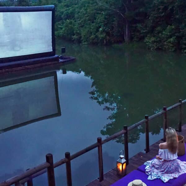 A woman sits on a wooden dock by a lake at dusk, facing an inflated movie screen floating on the water. Lanterns line the dock, and trees surround the lake, creating a serene outdoor cinema setting. This exclusive Soneva experience in Thailand is tranquil and illuminated by gentle lighting.