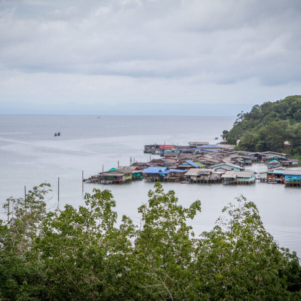 A coastal village with houses on stilts over water, surrounded by lush green trees. In the background, a vast body of water extends to the horizon under a cloudy sky, with small boats visible in the distance. This Thailand exclusive experience offers a landscape that appears serene and tranquil.