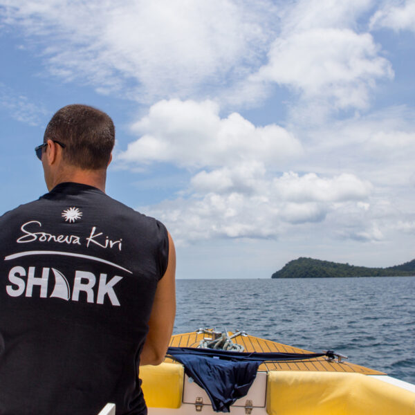 A person wearing a black sleeveless shirt with &quot;Soneva Kiri SHARK&quot; written on the back sits at the front of a yellow boat, looking out towards the open ocean. The sky is partly cloudy, and an island with lush greenery is visible in the distance. This Thailand exclusive experience truly embodies paradise.