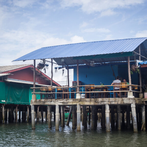 Colorful stilt houses with blue and red roofs sit above water in this exclusive Soneva experience in Thailand. Wooden railings, tables, and chairs adorn the porch of one house. The scene unfolds under a partly cloudy sky, with the structures supported by sturdy wooden pillars.