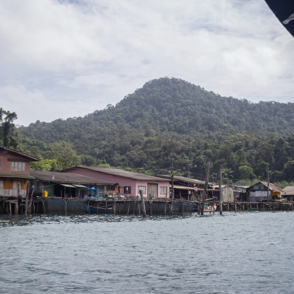 Nestled in a coastal village, exclusive waterside houses on stilts line the shore, framed by lush, green hills and an overcast sky. Some boats float near the houses on calm waters with gentle ripples. This serene setting offers a unique Thailand Soneva experience.