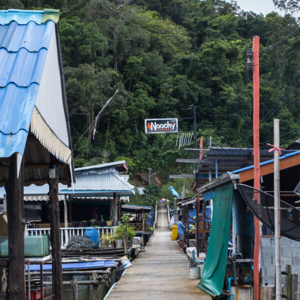 A narrow path runs through a fishing village with houses on stilts, leading towards a lush green hillside. A large sign reading &quot;Noochy&quot; is visible in the background, hanging above the trees. The scene is calm and overcast, offering a Thailand exclusive experience reminiscent of Soneva&#039;s tranquil retreats.