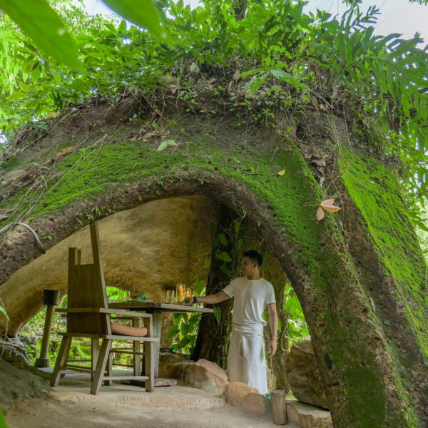 A man stands beside a rustic wooden table and chairs set under a large, moss-covered rock arch in a dense, lush forest. This Thailand exclusive experience offers a serene, green backdrop with sunlight and shade filtering through the trees, epitomizing the luxury of Soneva&#039;s natural retreats.