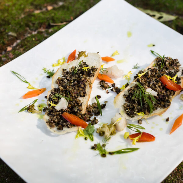 A white rectangular plate holds an artfully arranged dish featuring a bed of chopped greens and grains, garnished with thin carrot slices, leafy greens, and small edible flowers. This Thailand Exclusive Experience by Soneva is presented on a rough, natural surface with a mossy texture.